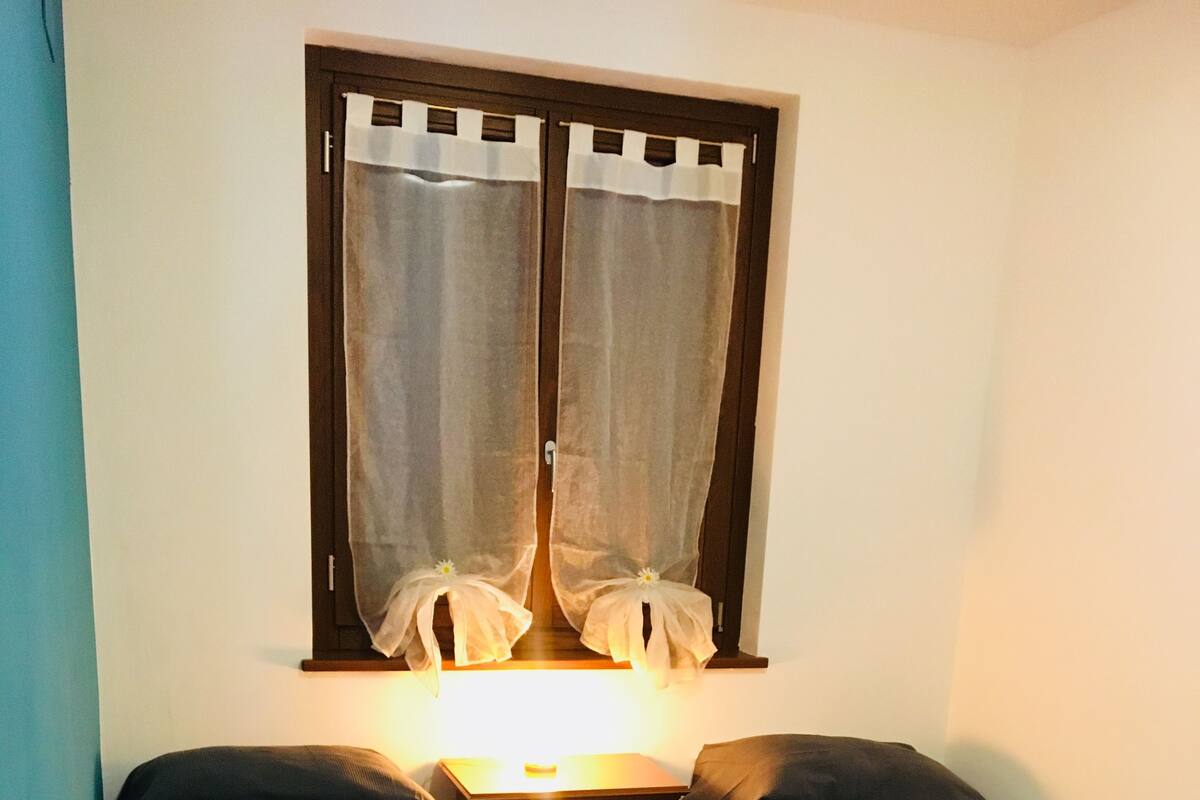 A small, cozy room with two single beds, a wooden floor, a nightstand with a lamp, and a window with curtains.