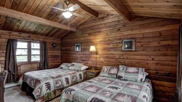 A cabin-style bedroom with two beds, wooden walls, a ceiling fan, and a window with curtains.