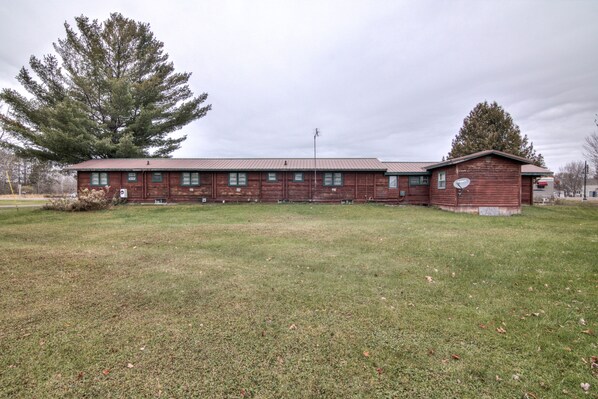 A long, single-story wooden building with a metal roof, surrounded by a grassy field and large trees.