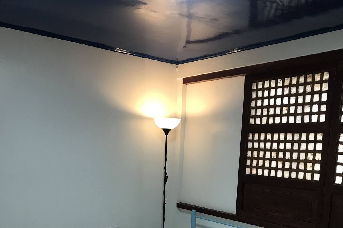 Ein Einzelbett mit Metallgestell und einer weißen Matratze in einem Zimmer mit weißen Wänden und einem Holzfenster mit Gitterstruktur.