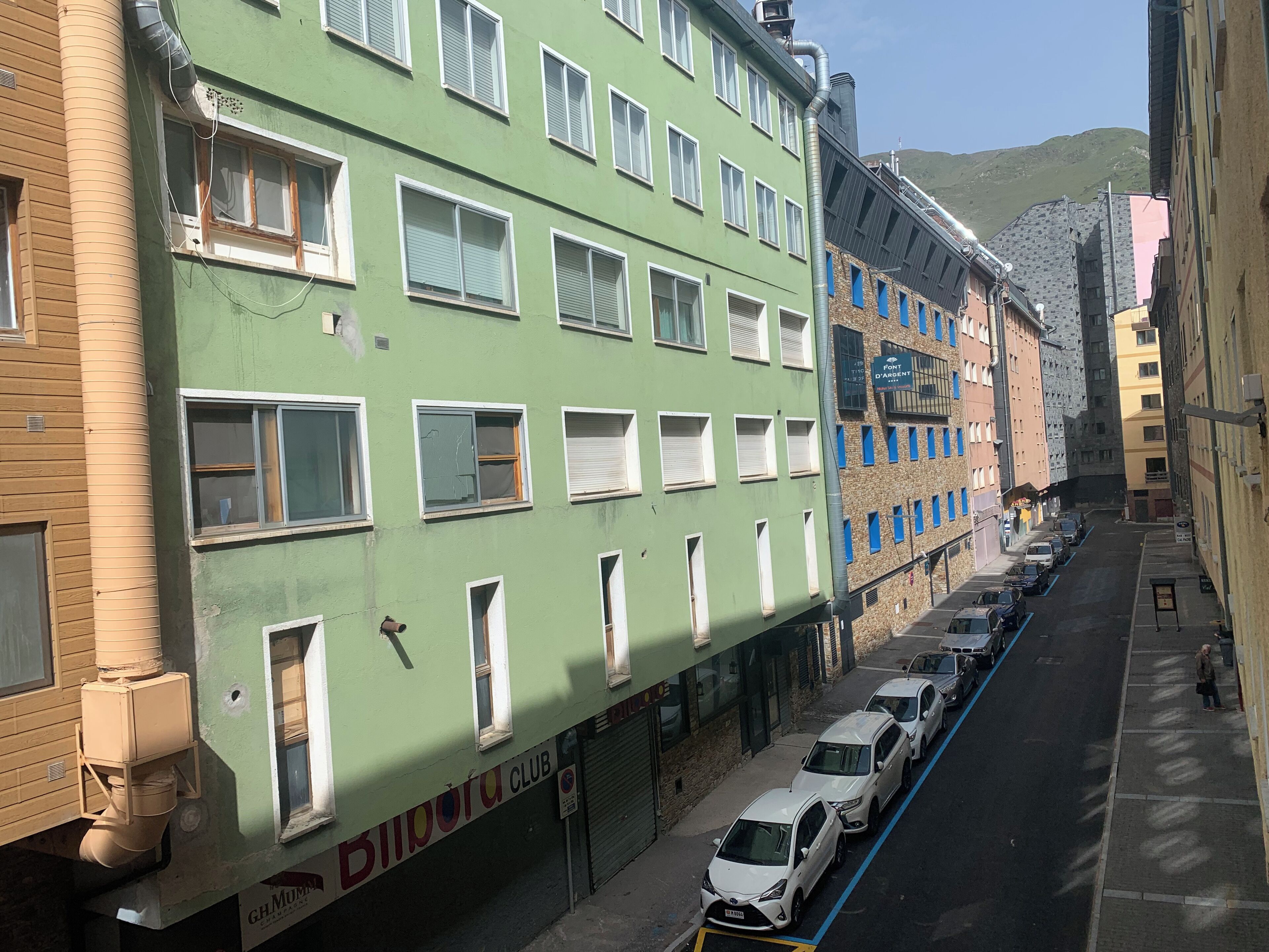 Una calle estrecha bordeada de edificios de varios pisos, algunos con señalización visible, coches aparcados y un cielo despejado.