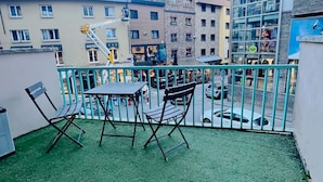 Un balcon avec deux chaises et une table, donnant sur une rue bordée d’immeubles et de boutiques.
