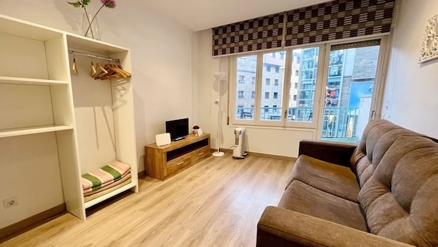 Un salon avec un plancher en bois, un canapé marron, un téléviseur posé sur un meuble en bois et une fenêtre avec des stores.