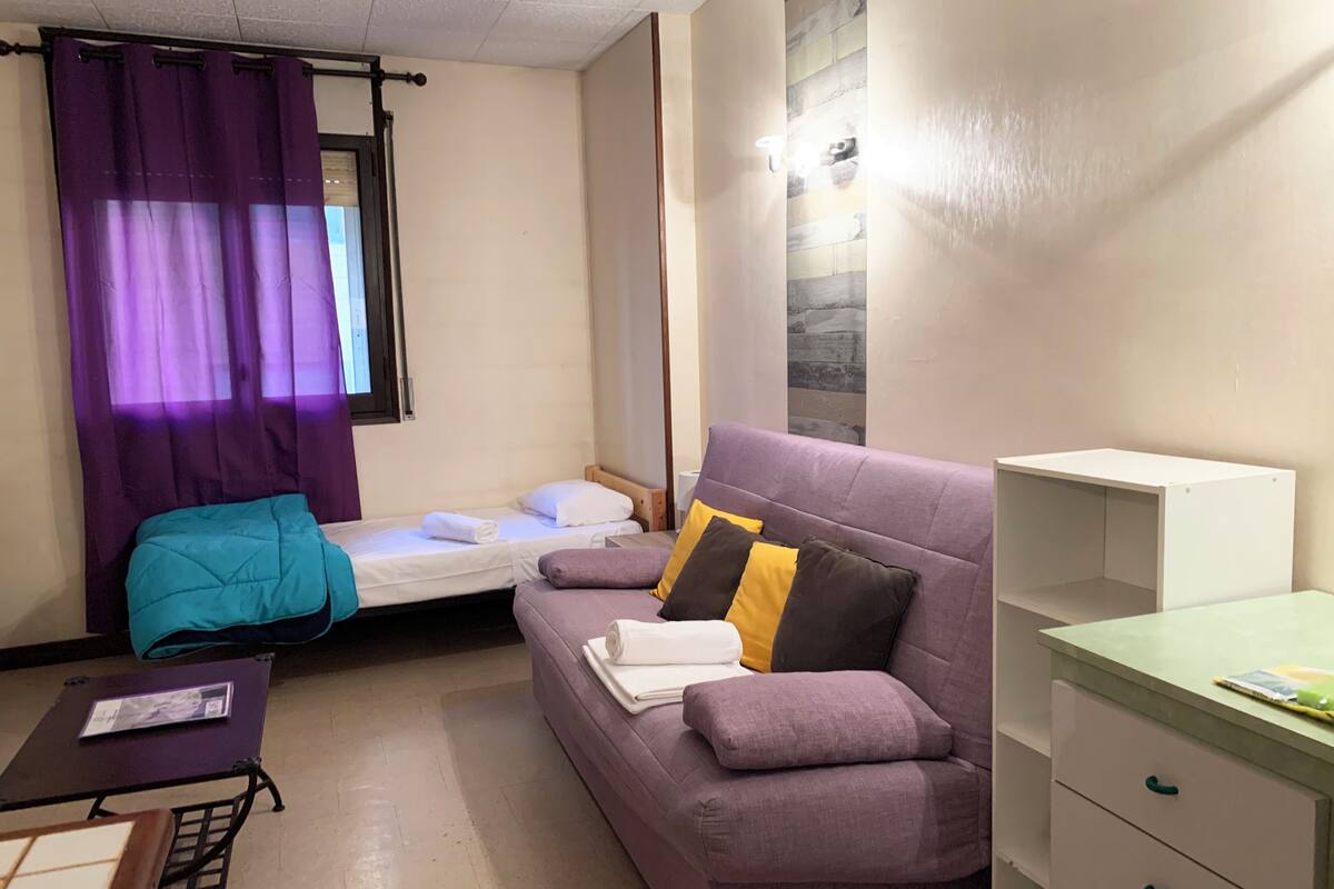 Una habitación pequeña y bien iluminada con un sofá morado, una cama con ropa blanca, una mesita con una revista y un escritorio verde con cajones.