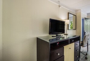 Habitación de hotel con televisor de pantalla plana sobre un mueble negro y beige, un espejo encima y una silla al fondo.