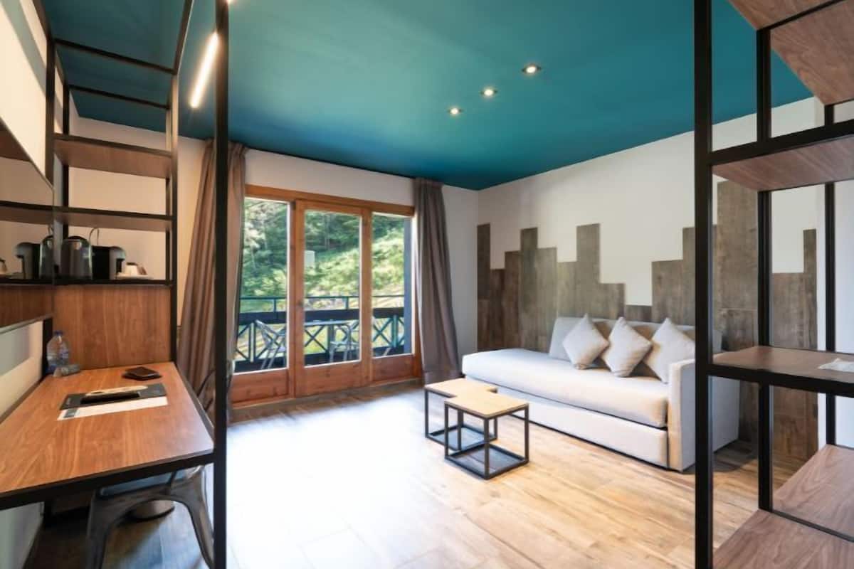 Een moderne kamer met een houten bureau, een witte bank en een balkon met uitzicht op groen.