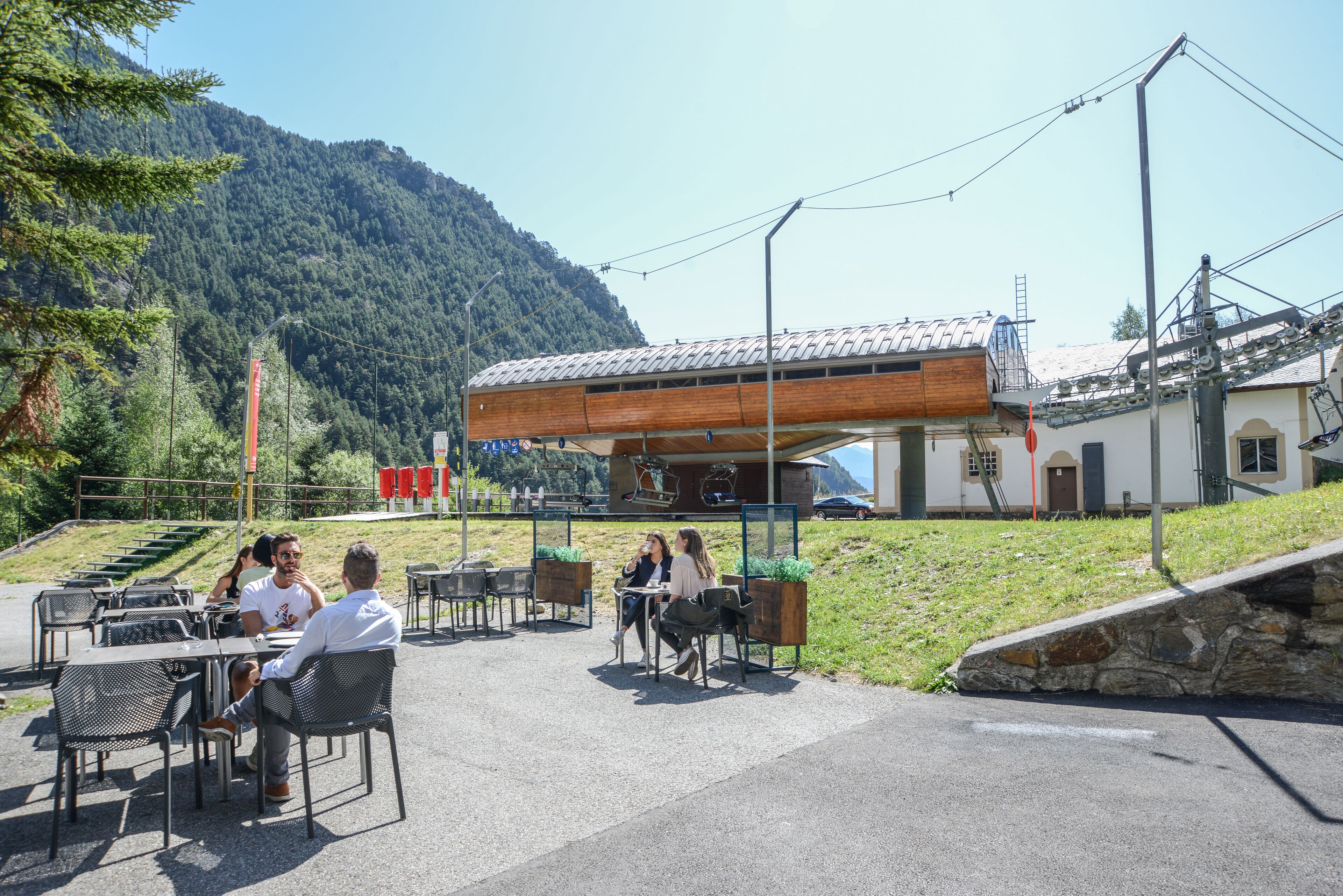 An outdoor seating area with tables and chairs, a building with a sloped roof, and a mountainous backdrop.