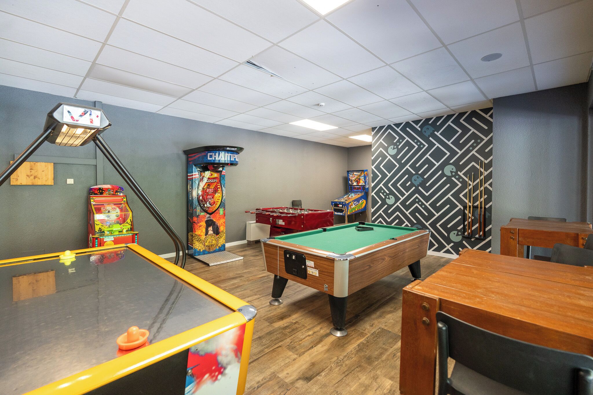 A pool table, foosball table, and arcade games in a room with wooden flooring and patterned walls.