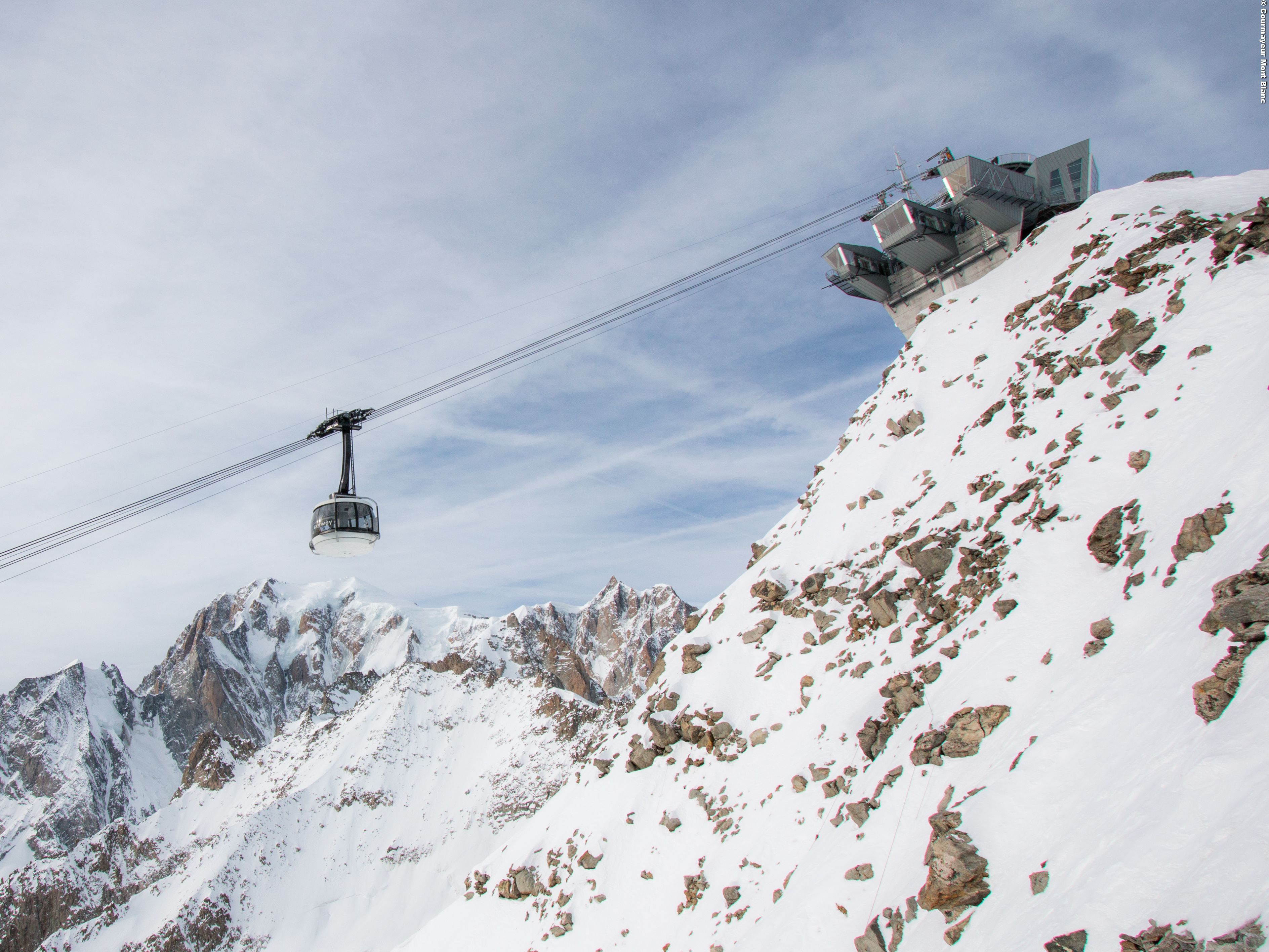 Eine Seilbahn in einer gebirgigen Landschaft mit felsigem Gelände und schneebedeckten Gipfeln.