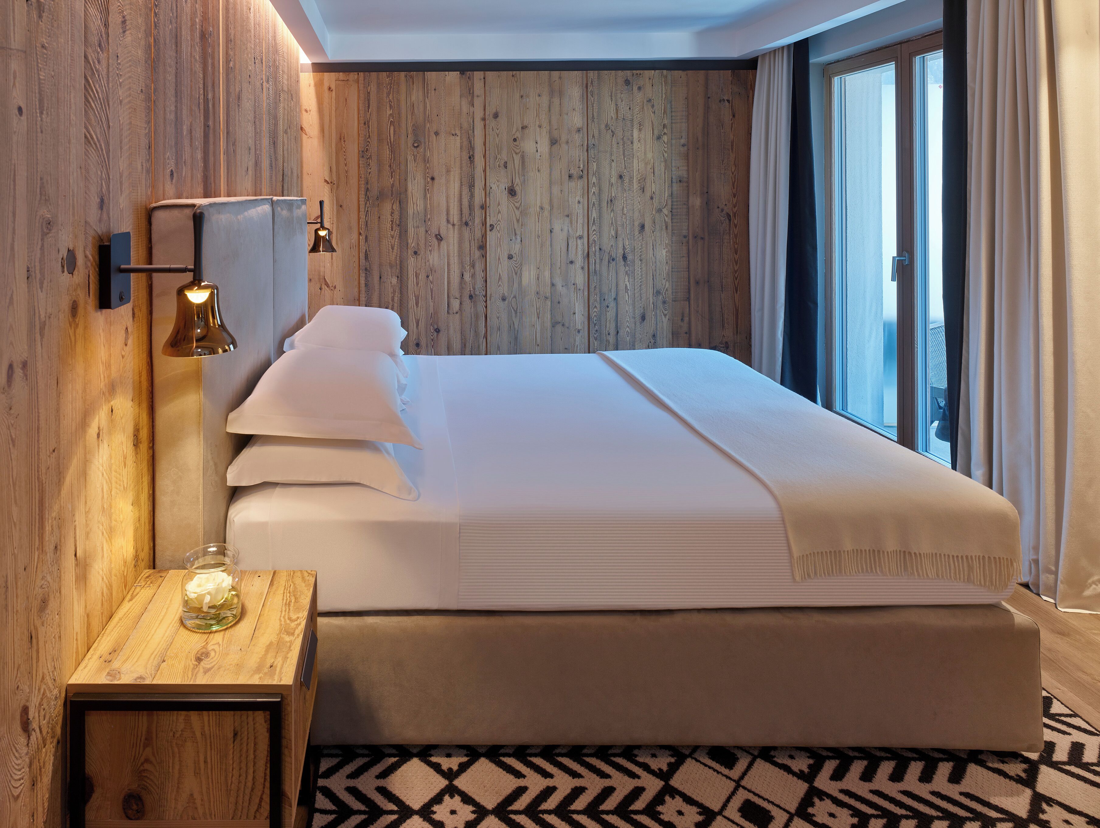 A neatly made bed with white linens, a wooden headboard, a bedside table with a lamp, and a view of the outdoors through a window.