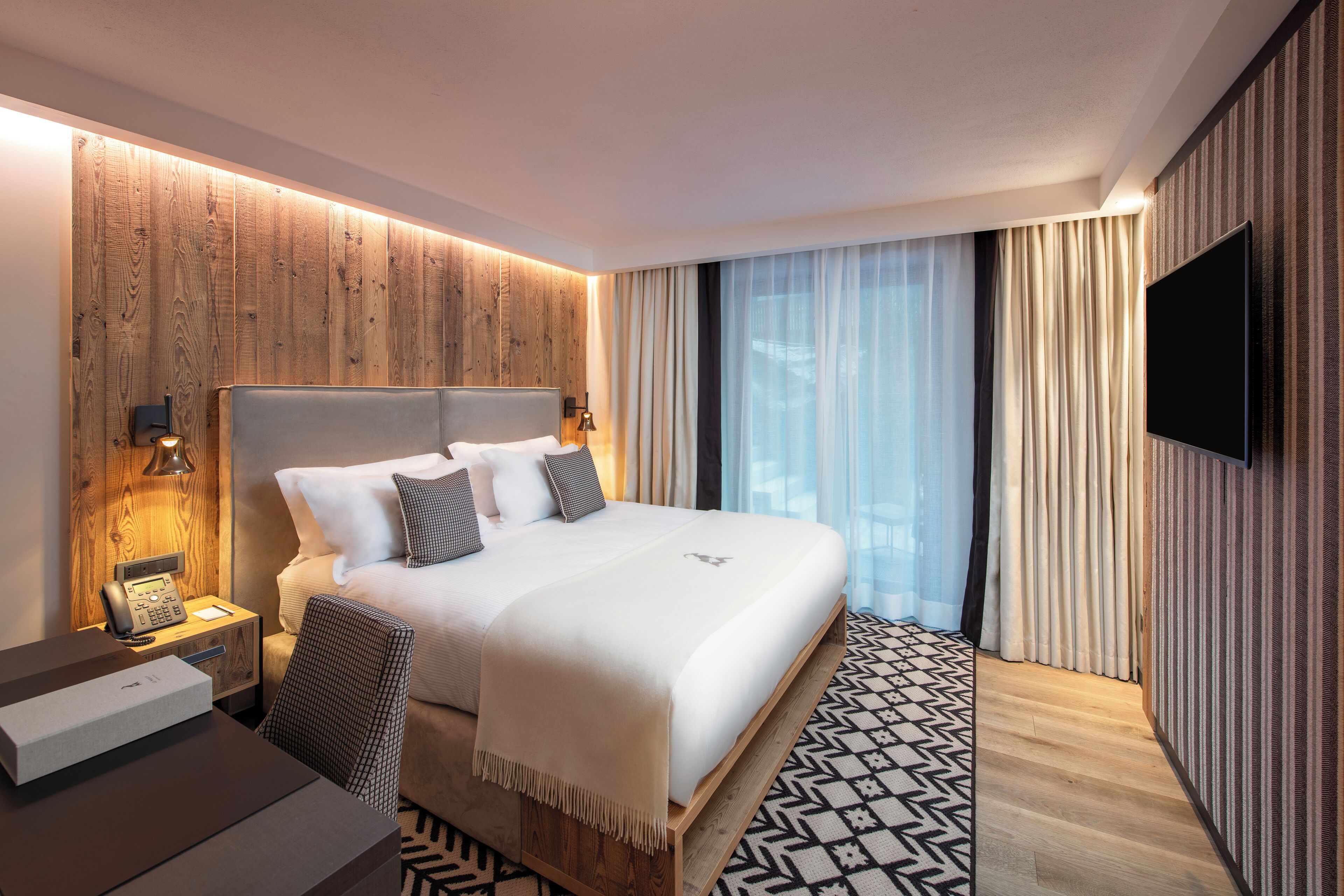 Een moderne hotelkamer met een groot bed, een nachtkastje met een lamp, een flatscreen televisie aan de muur en een kamerkleed met een patroon.