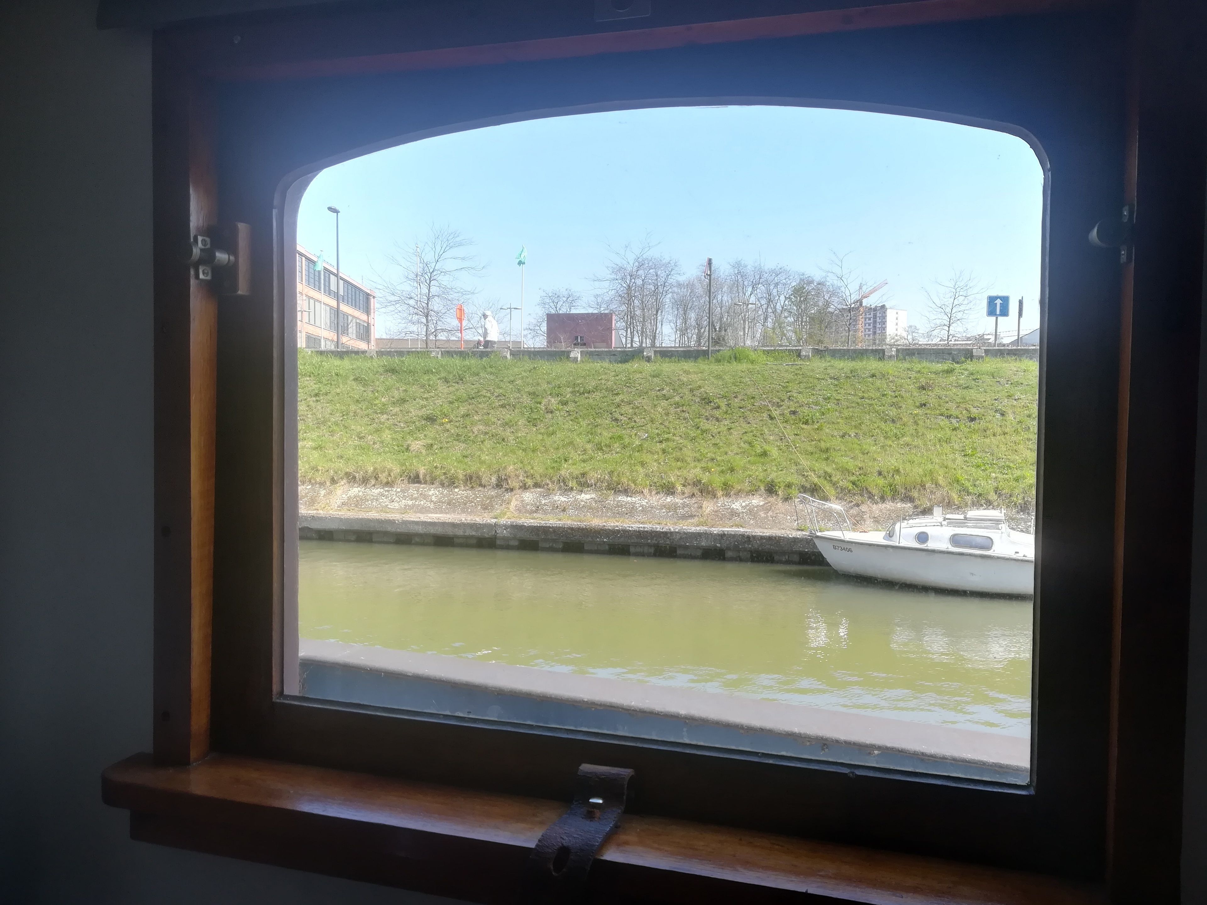 Blick aus einem Schiffsfenster auf einen Fluss, mit einer grünen Uferböschung und Gebäuden im Hintergrund.