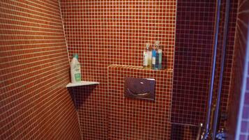 A small bathroom with a toilet, a shower, and a shelf with toiletries.
