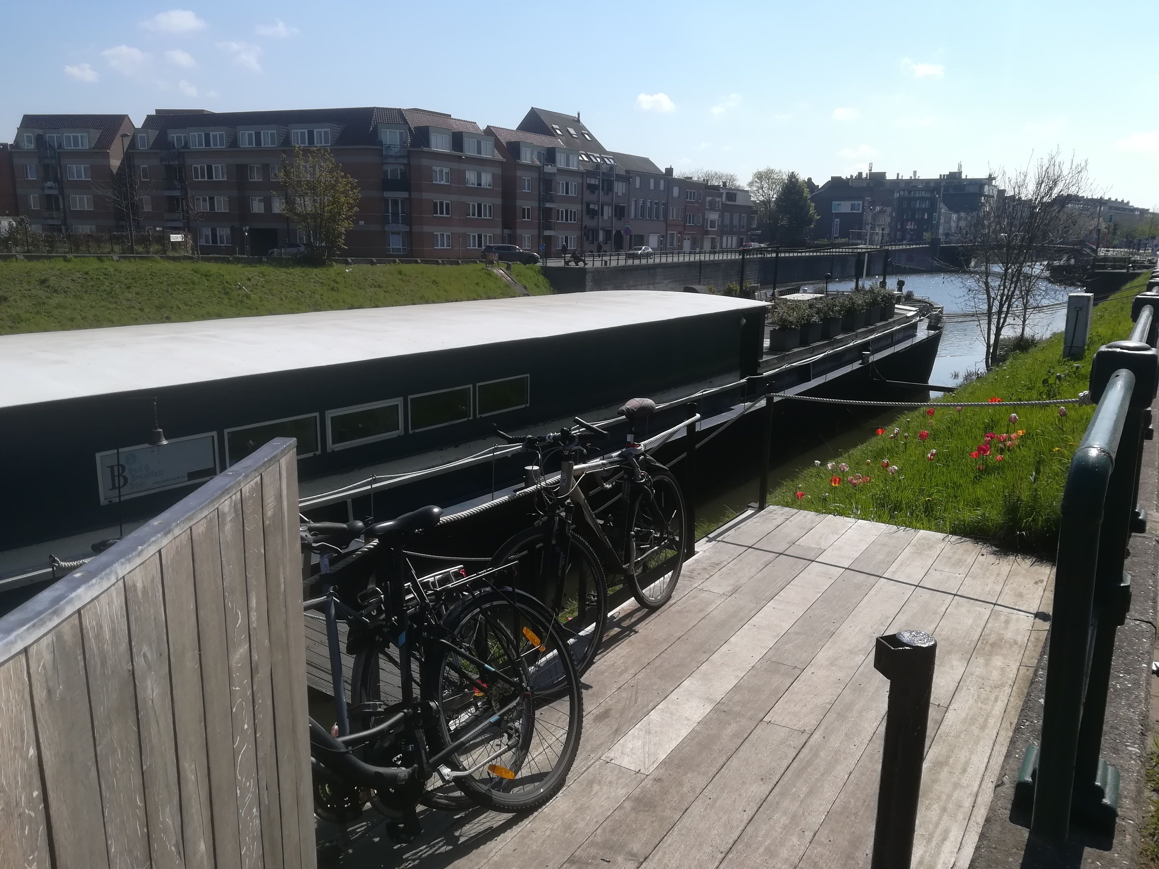 Ein Uferweg mit abgestellten Fahrrädern an der Brüstung, eine Brücke im Hintergrund und Wohnhäuser am gegenüberliegenden Ufer.
