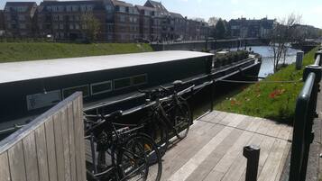 Ein Uferweg mit abgestellten Fahrrädern an der Brüstung, eine Brücke im Hintergrund und Wohnhäuser am gegenüberliegenden Ufer.