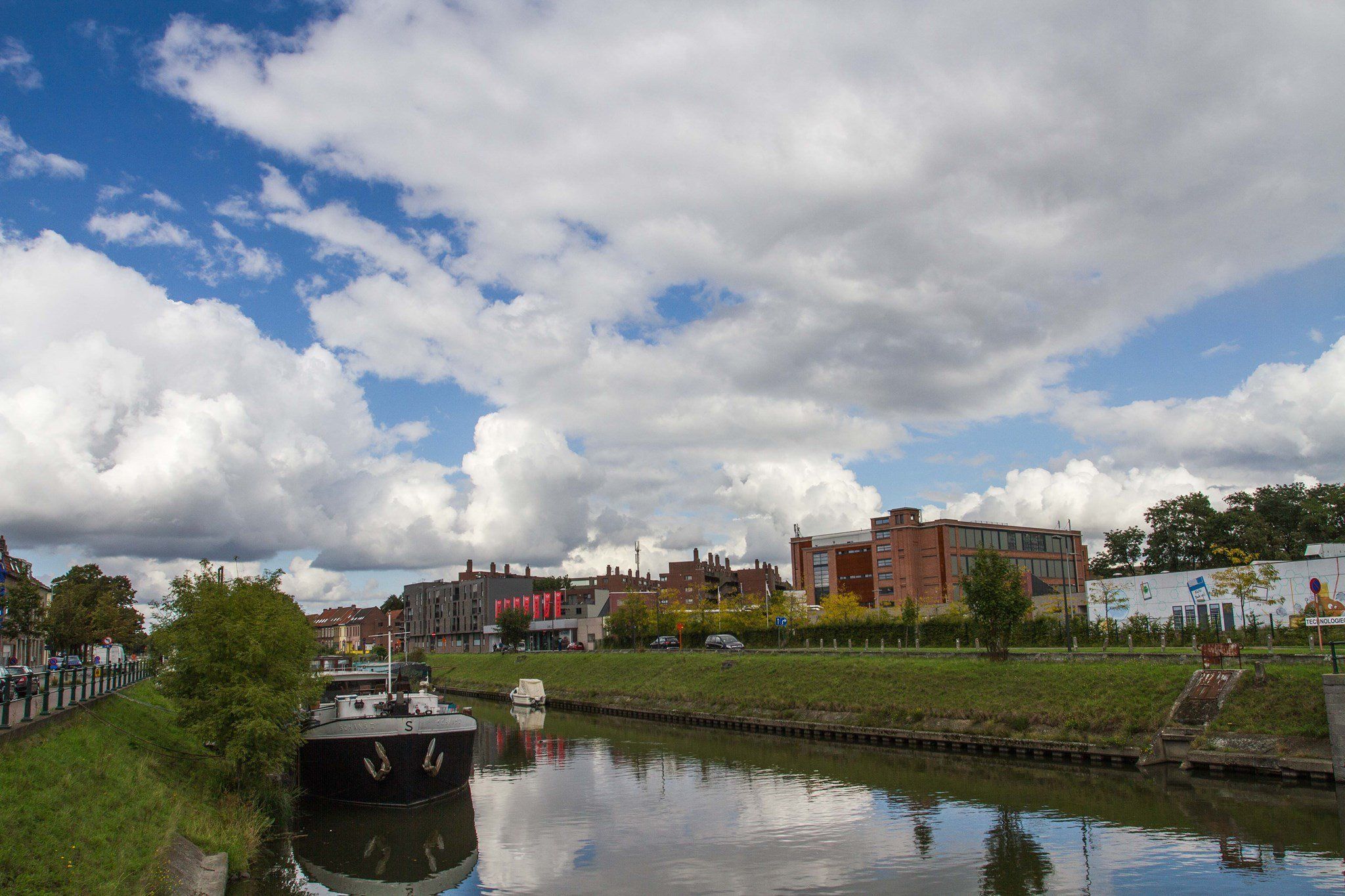 Un canal avec un bateau amarré sur la rive gauche, des bâtiments en arrière-plan et un ciel partiellement nuageux.