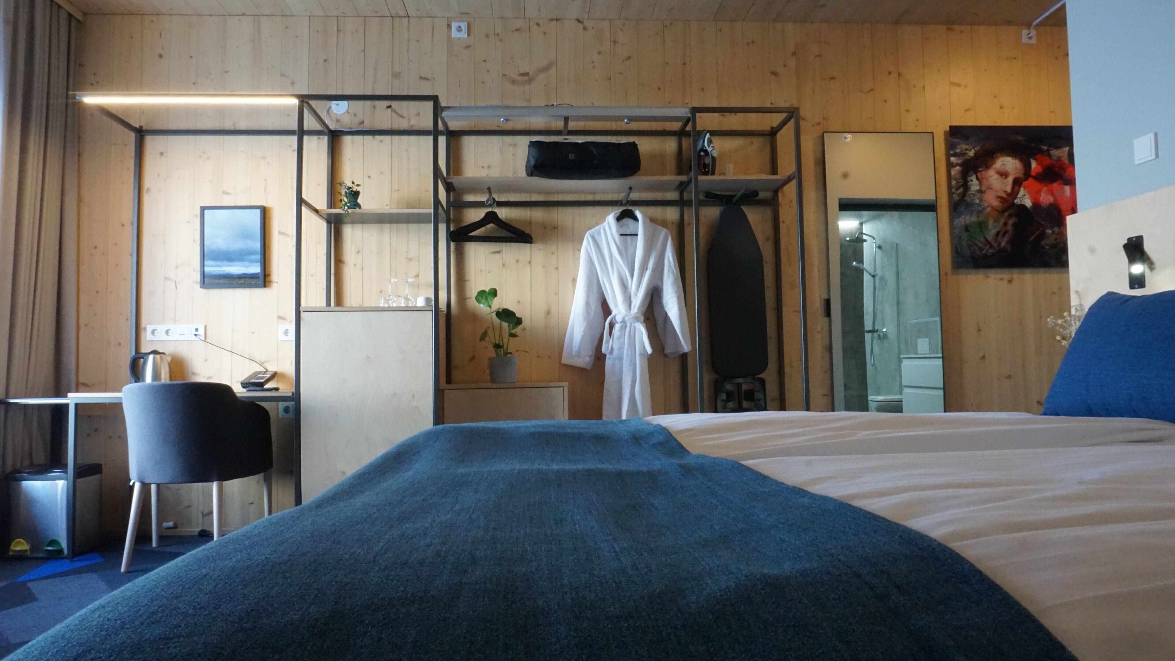 Une chambre d’hôtel avec un lit, un bureau, une chaise, une robe accrochée, un espace douche et un tableau au mur.