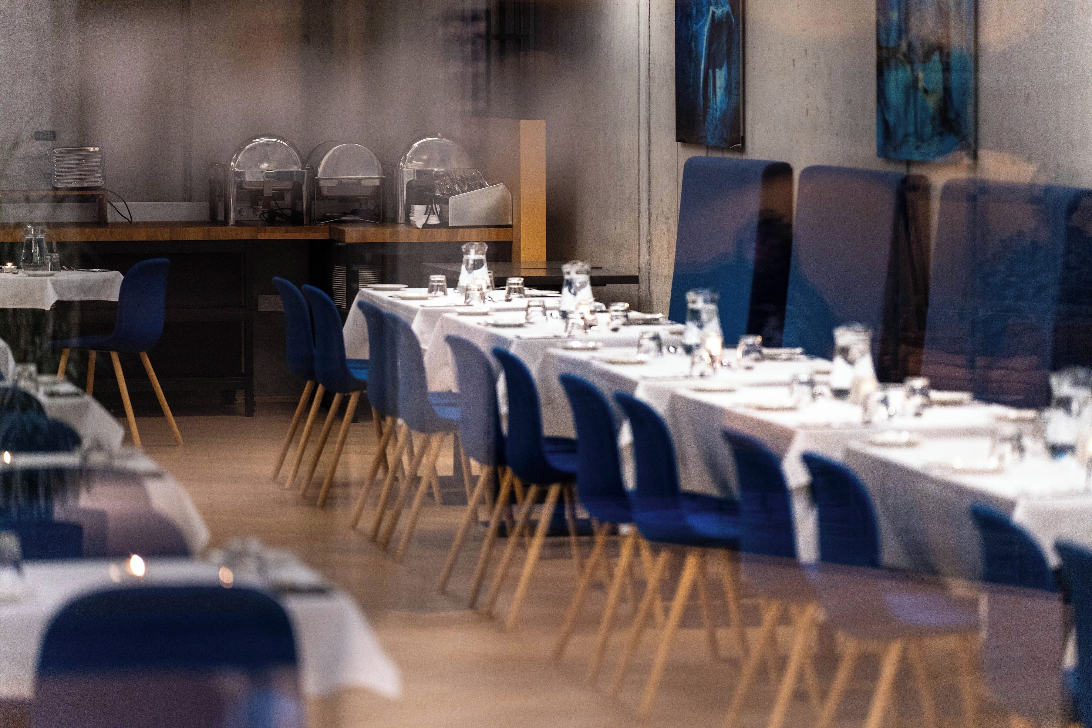 Un restaurante con mesas largas dispuestas para comer, sillas azules y una barra con equipo para servicio de alimentos.