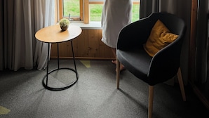 A room with a large window, a wooden floor, a small round table with a plant, and a black chair with a yellow cushion.