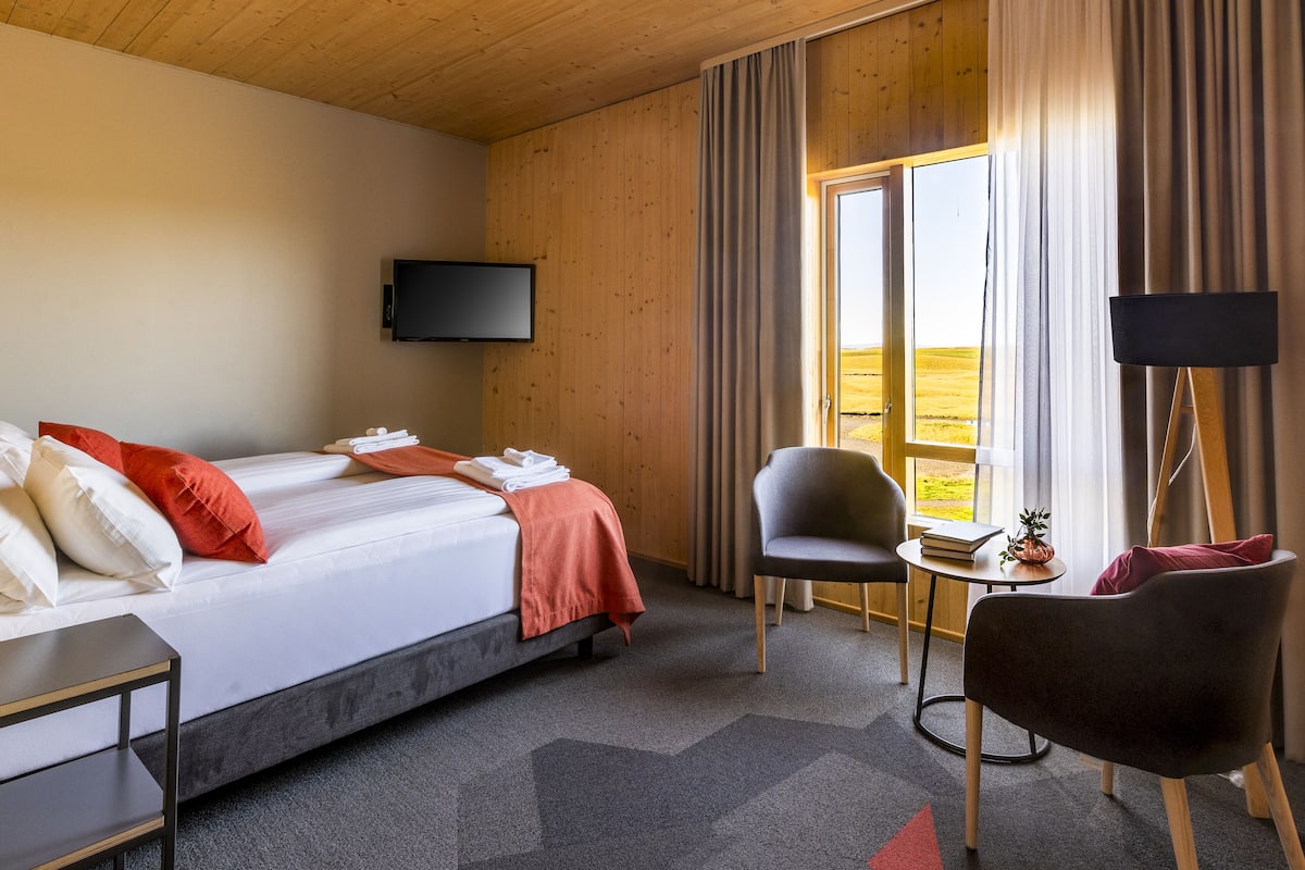 A hotel room with a bed, a chair, a small table with a book and a pineapple, and a TV mounted on the wall.