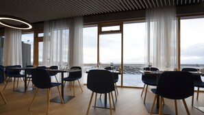 A dining area with a view of the sea, featuring a table set with wine glasses and plates, and blue chairs with wooden legs.