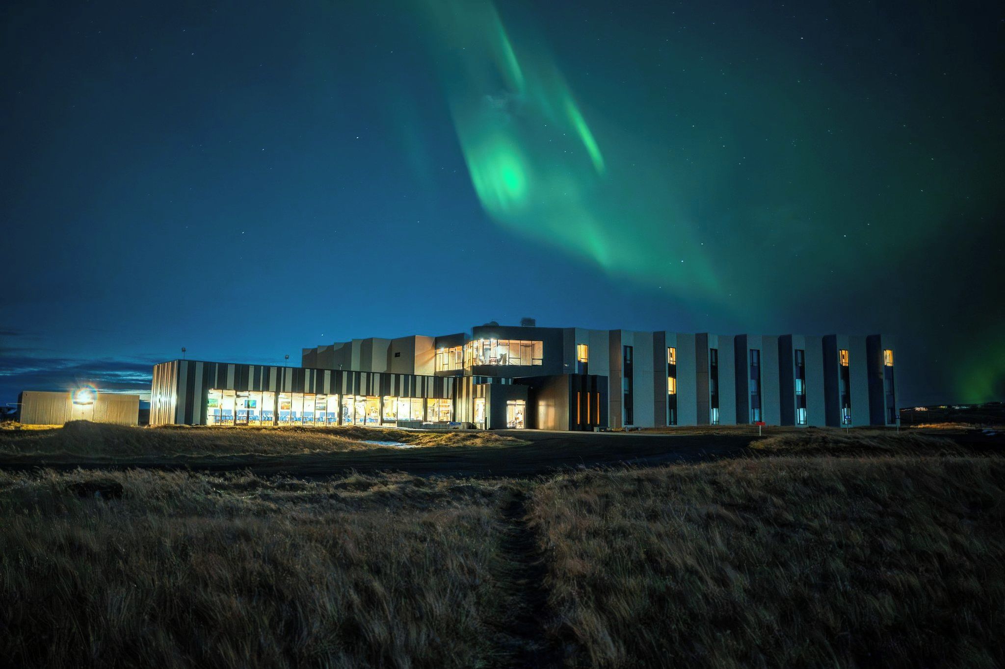 A modern building with a flat roof and large windows, located in a grassy area with a view of the night sky and northern lights.
