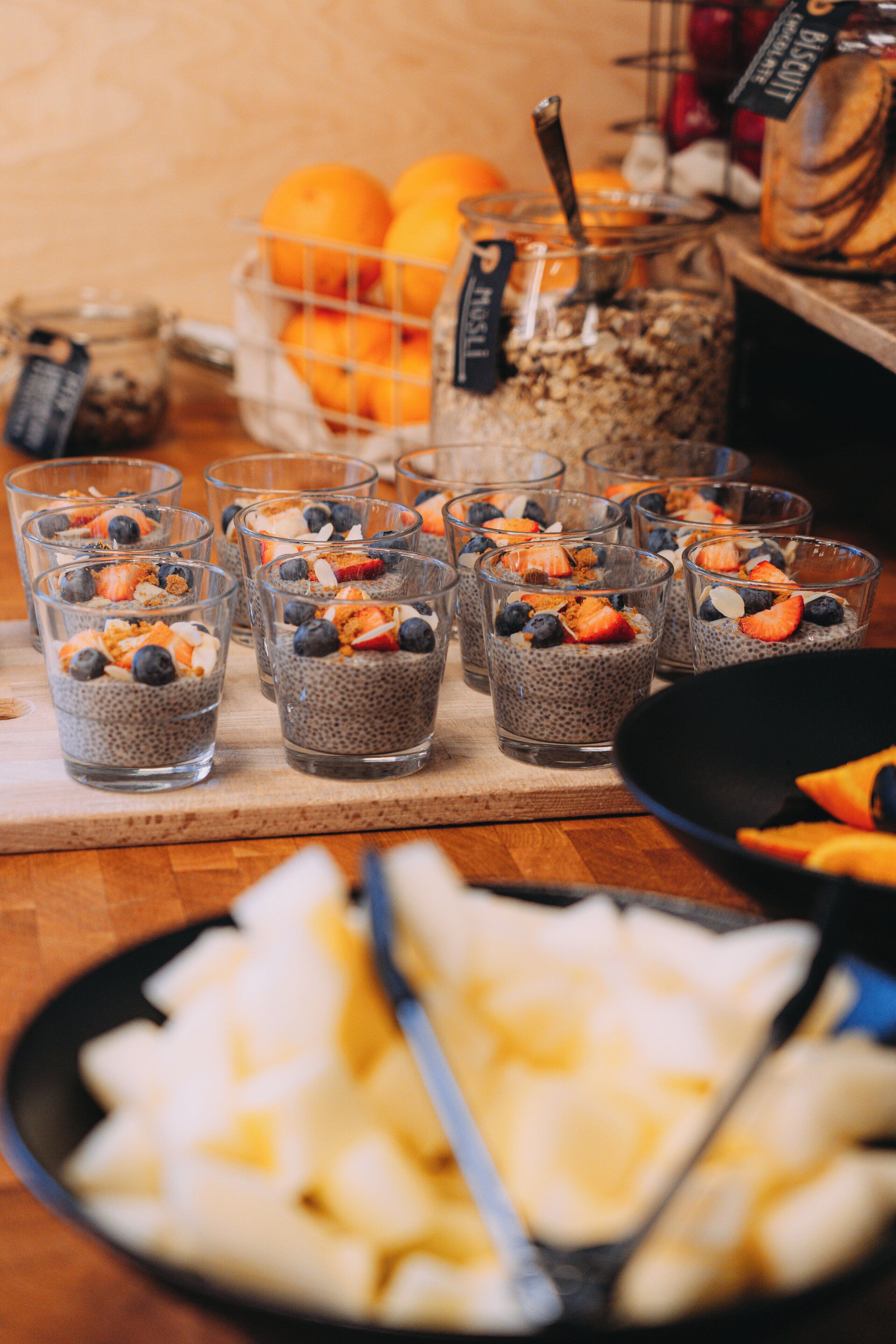 A table with chia seed pudding cups topped with fruits and granola, a basket of oranges, and a bowl of sliced apples.