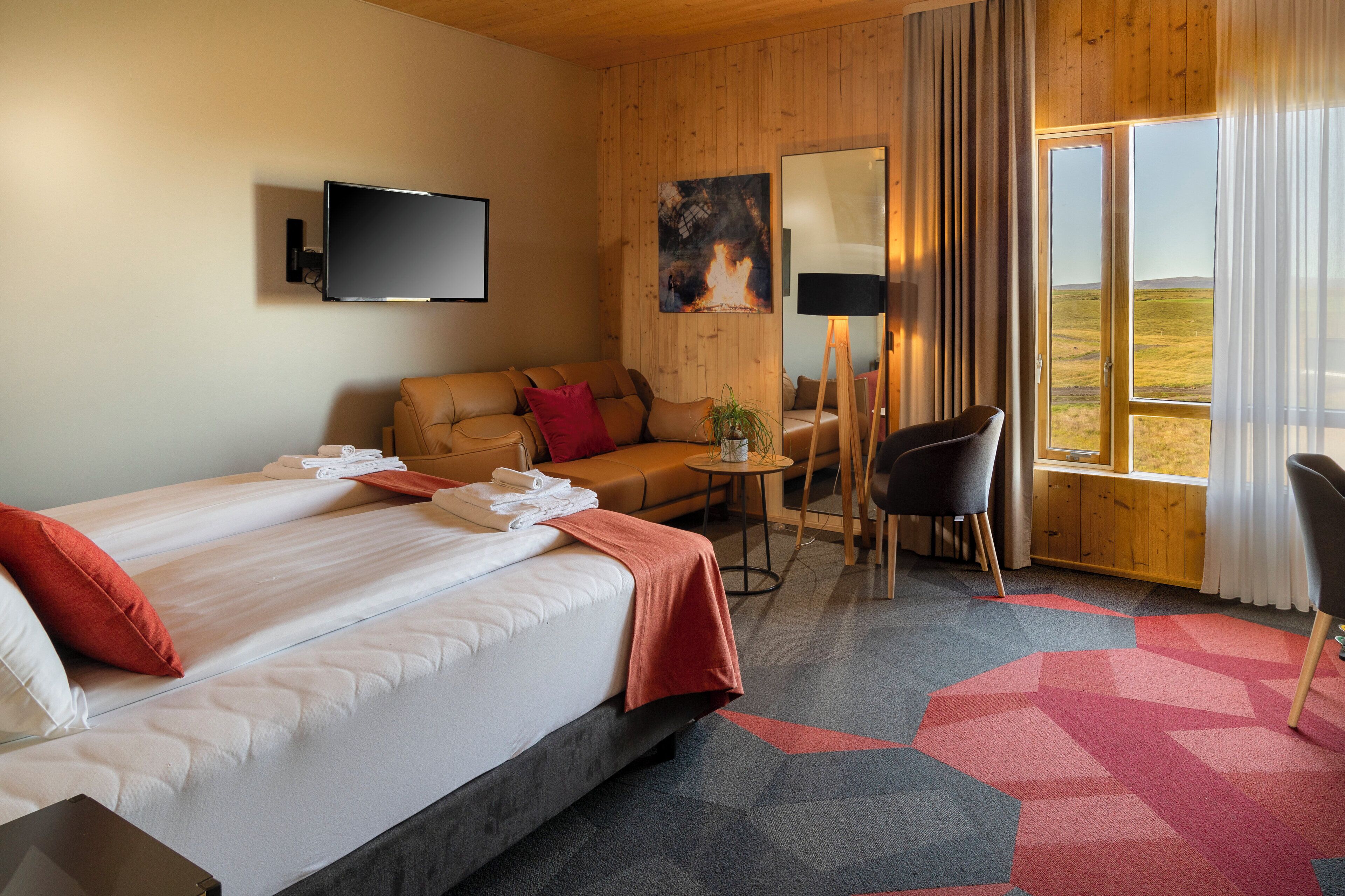 Habitación de hotel con una cama grande, un sofá, un escritorio y una silla. Hay un televisor montado en la pared y una ventana con cortinas.