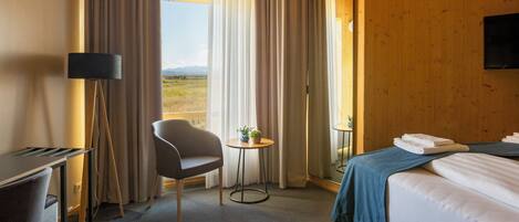 A hotel room with a bed, a desk, a chair, a small table with a plant, and a view of the outdoors.