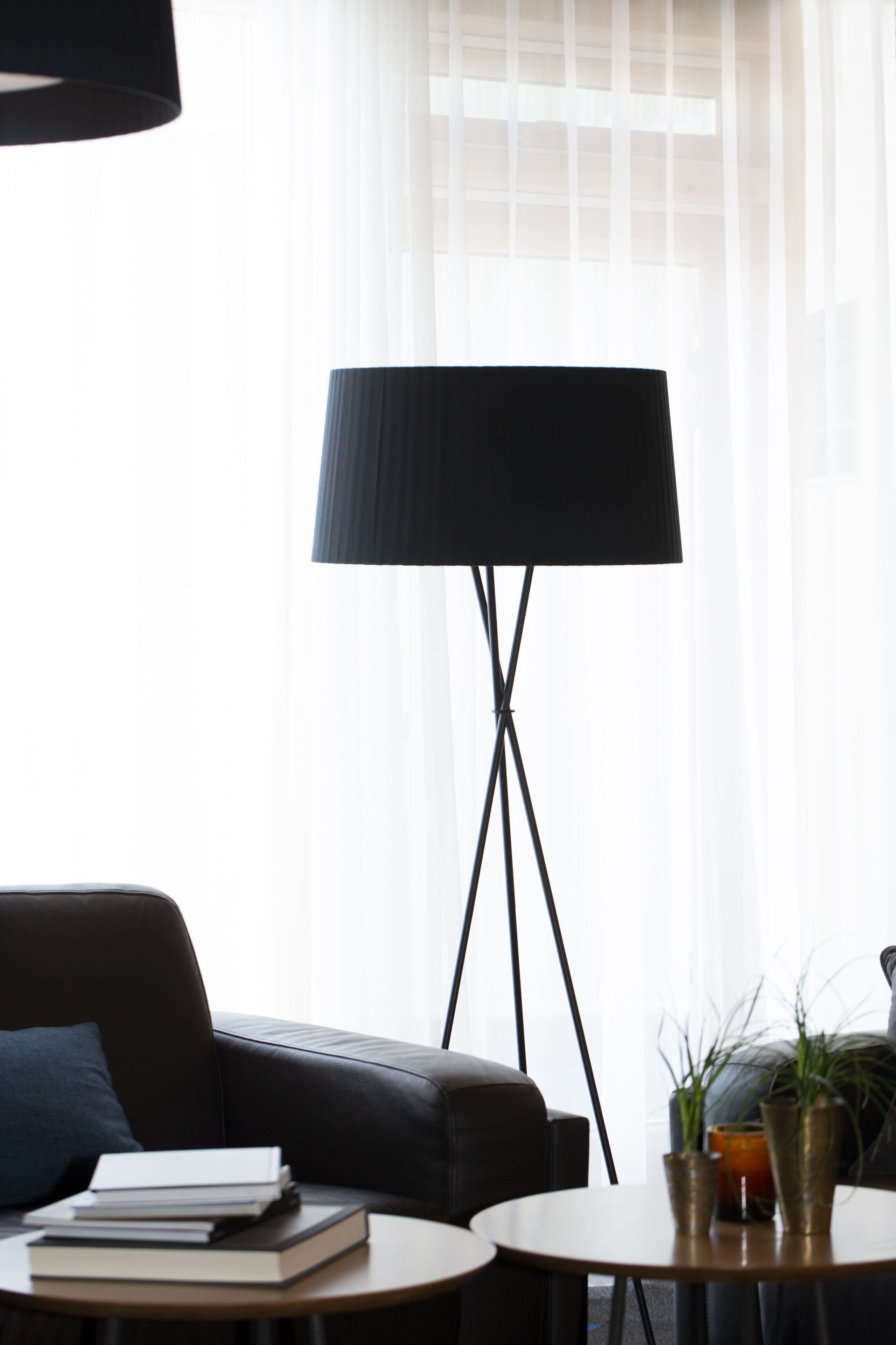 A modern living room with a black leather sofa, a round wooden coffee table, and a floor lamp with a black shade. There are books stacked on the table and two potted plants on the floor.