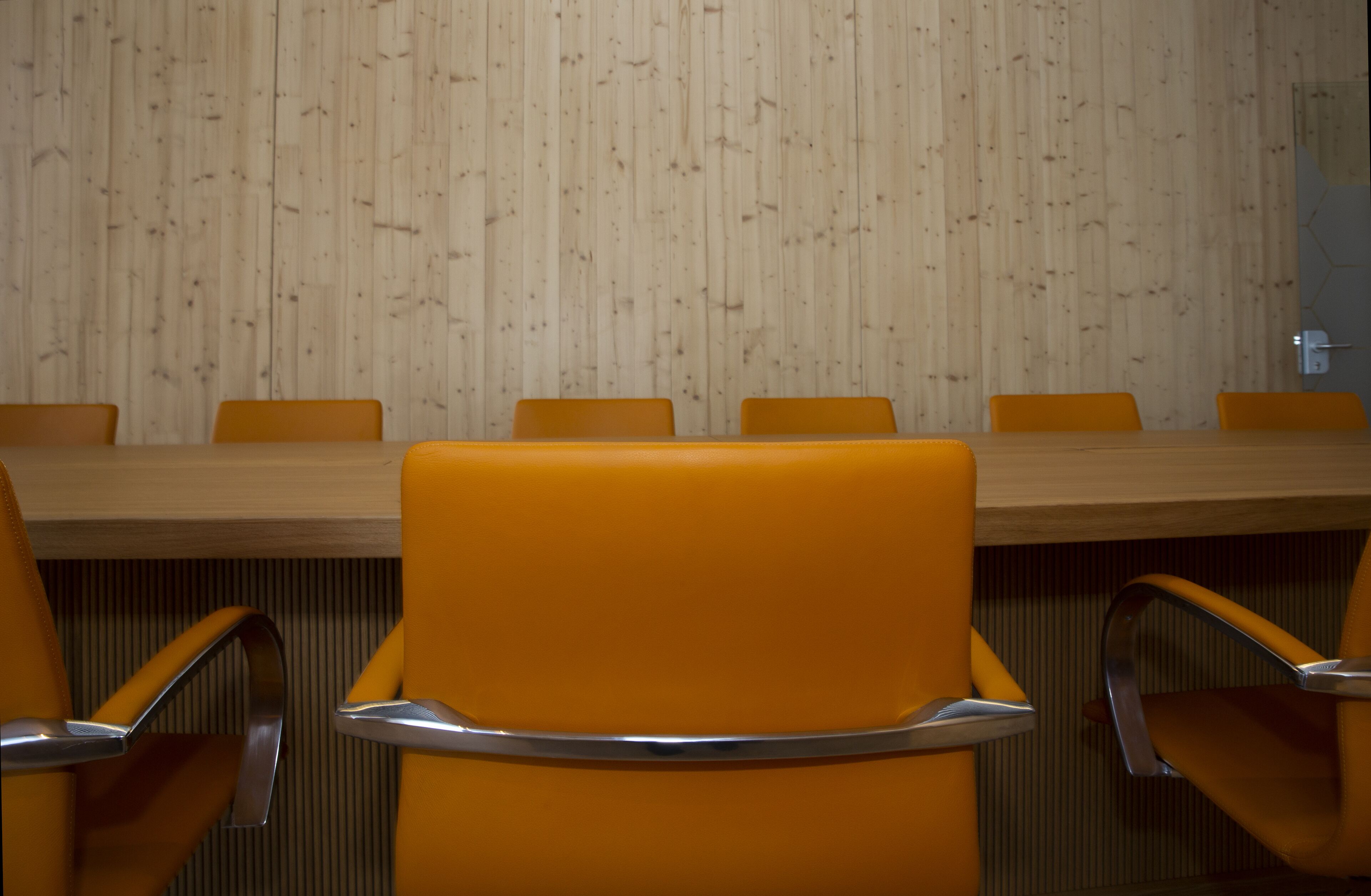 Un salón de conferencias con pared de paneles de madera, una mesa larga y sillas naranja con reposabrazos metálicos.