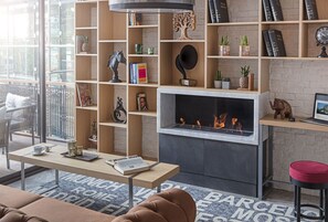 A modern living room with a fireplace, wooden shelves with books and decorative items, a dining table with a laptop, and a red chair. - TRYP by Wyndham Ankara Oran (Ankara)
