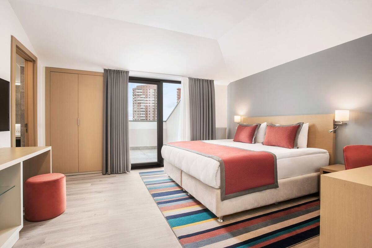 A hotel room with a bed, a desk, a chair, a TV, and a view of the city.