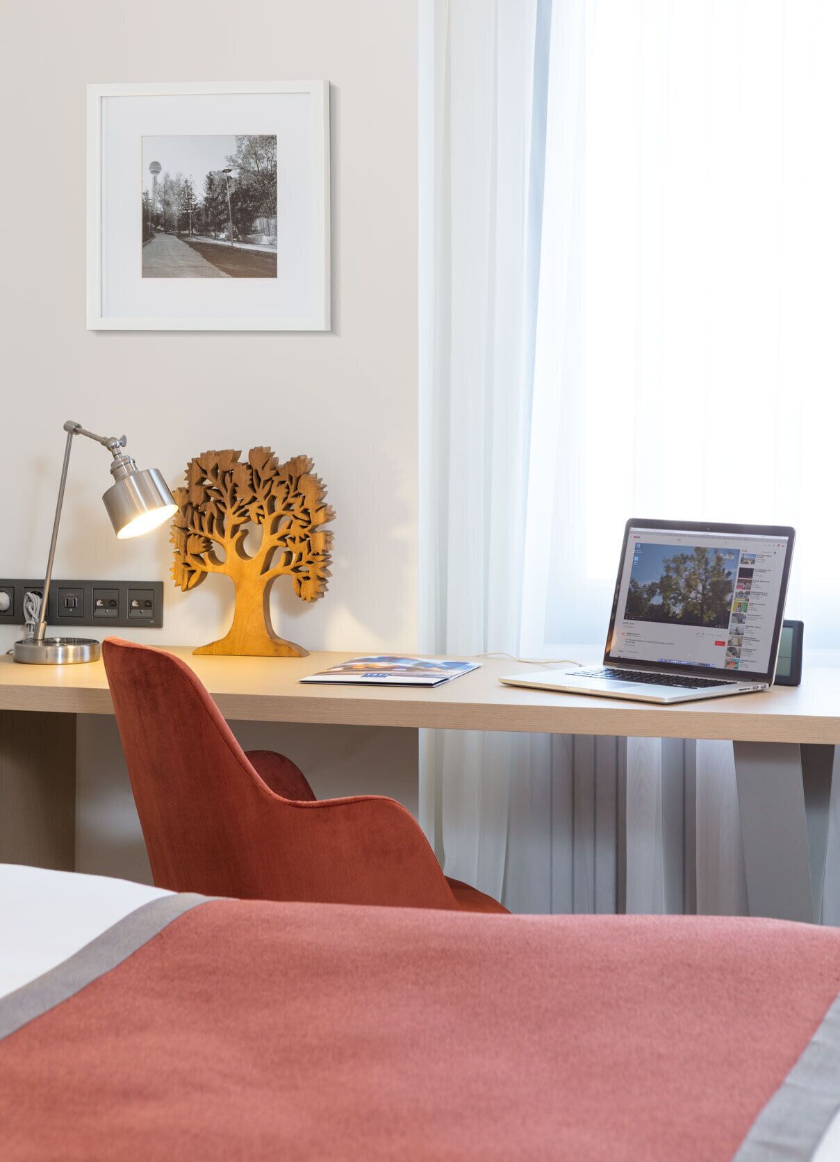 A modern hotel room with a wooden desk, a laptop, a lamp, and a framed picture on the wall.