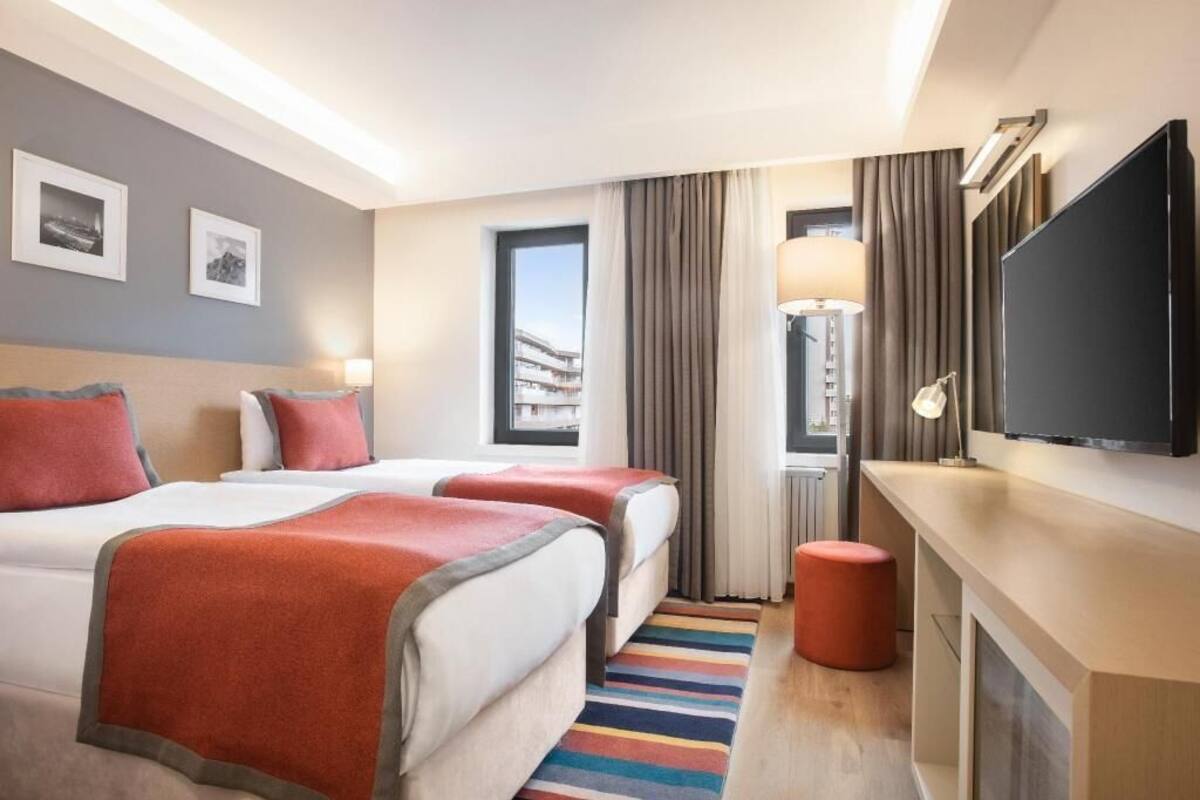 A hotel room with two beds, a TV, a lamp, and a view of the city.