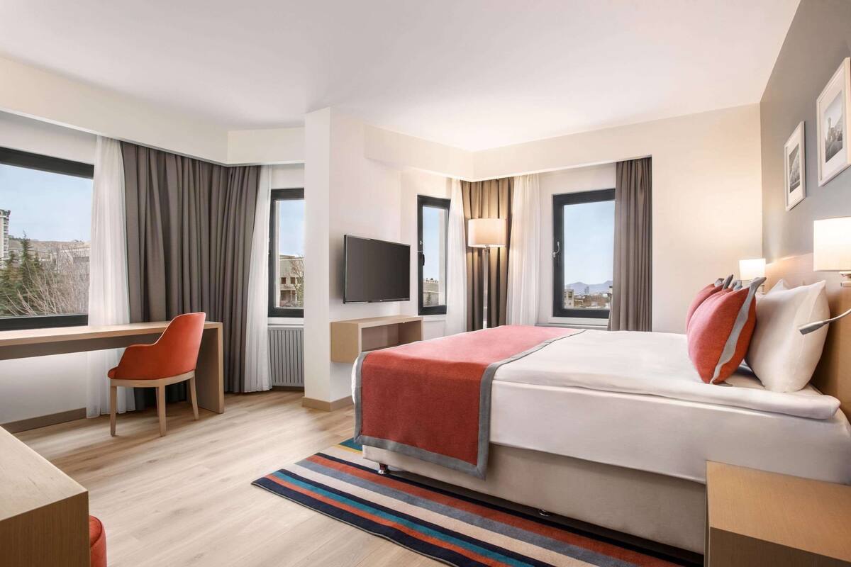 A hotel room with a bed, a desk, a chair, a TV, and a window with curtains.