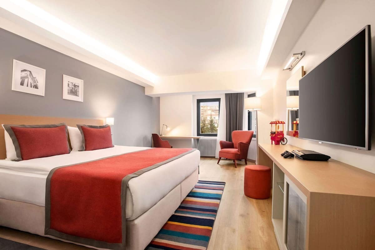 A hotel room with a bed, a desk with a TV, a chair, and a window.