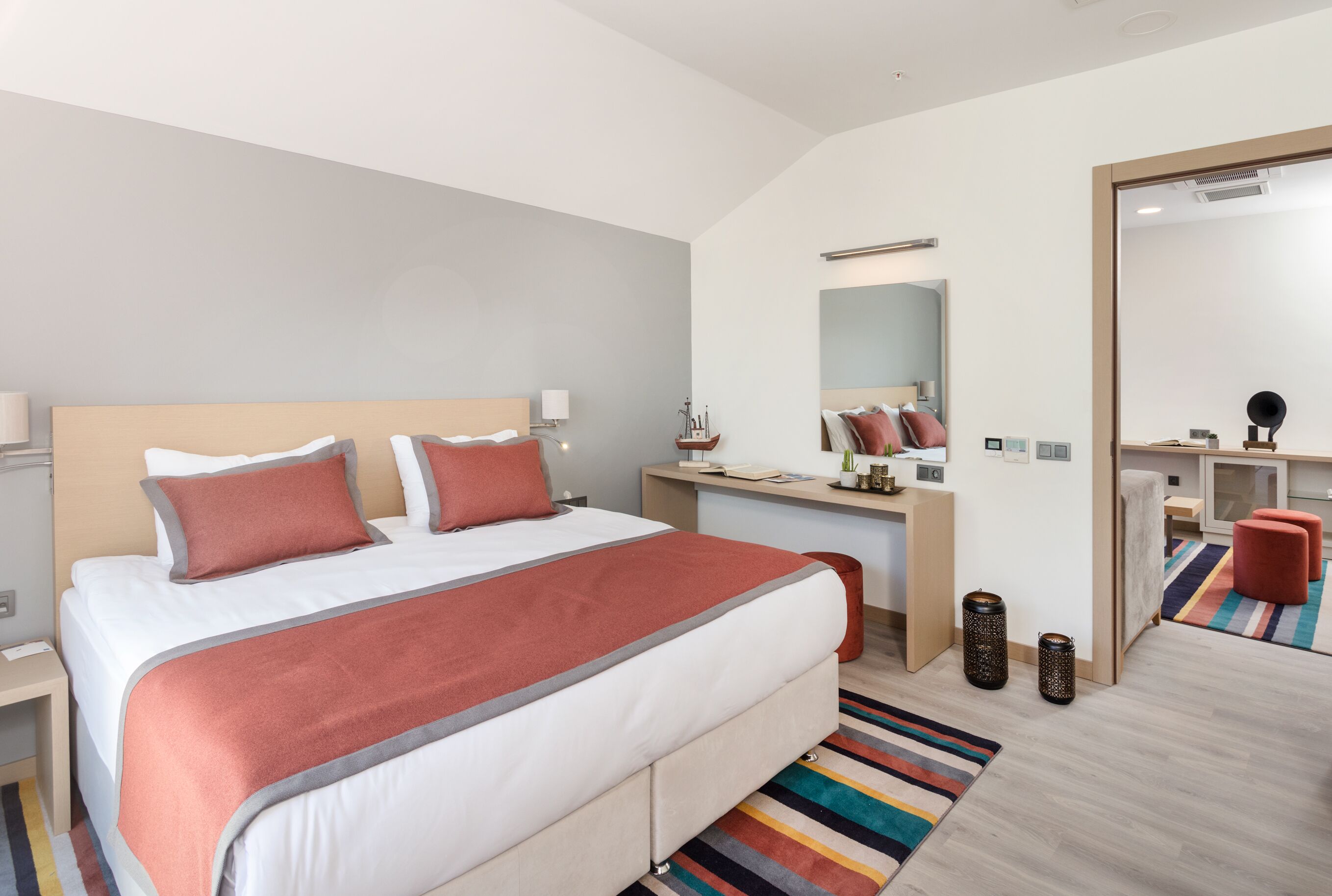Moderni hotellihuone, jossa on suuri sänky, työpöytä, peili ja värikäs raidallinen matto.