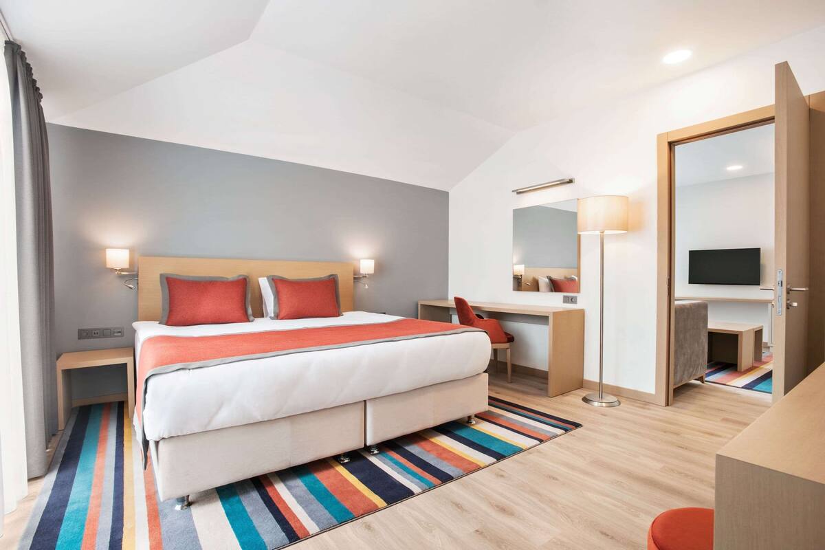 A modern hotel room with a large bed, a desk, a chair, a TV, and a mirror.