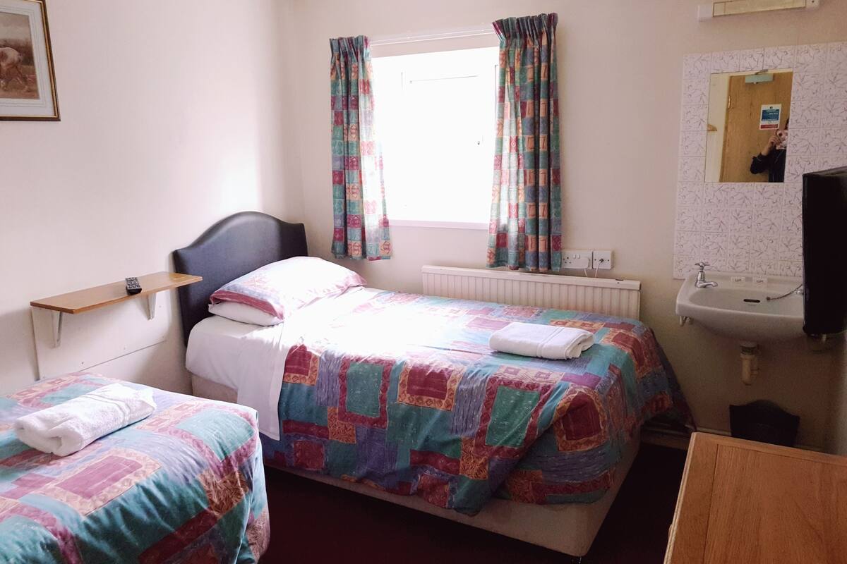 A hotel room with two beds, a window with curtains, a bedside table with a phone, a radiator, a sink, and a mirror.