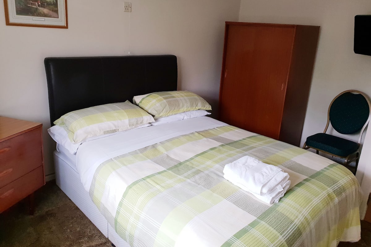 A neatly made bed with a checkered bedspread and pillows, a wooden wardrobe, a chair, and a painting on the wall.