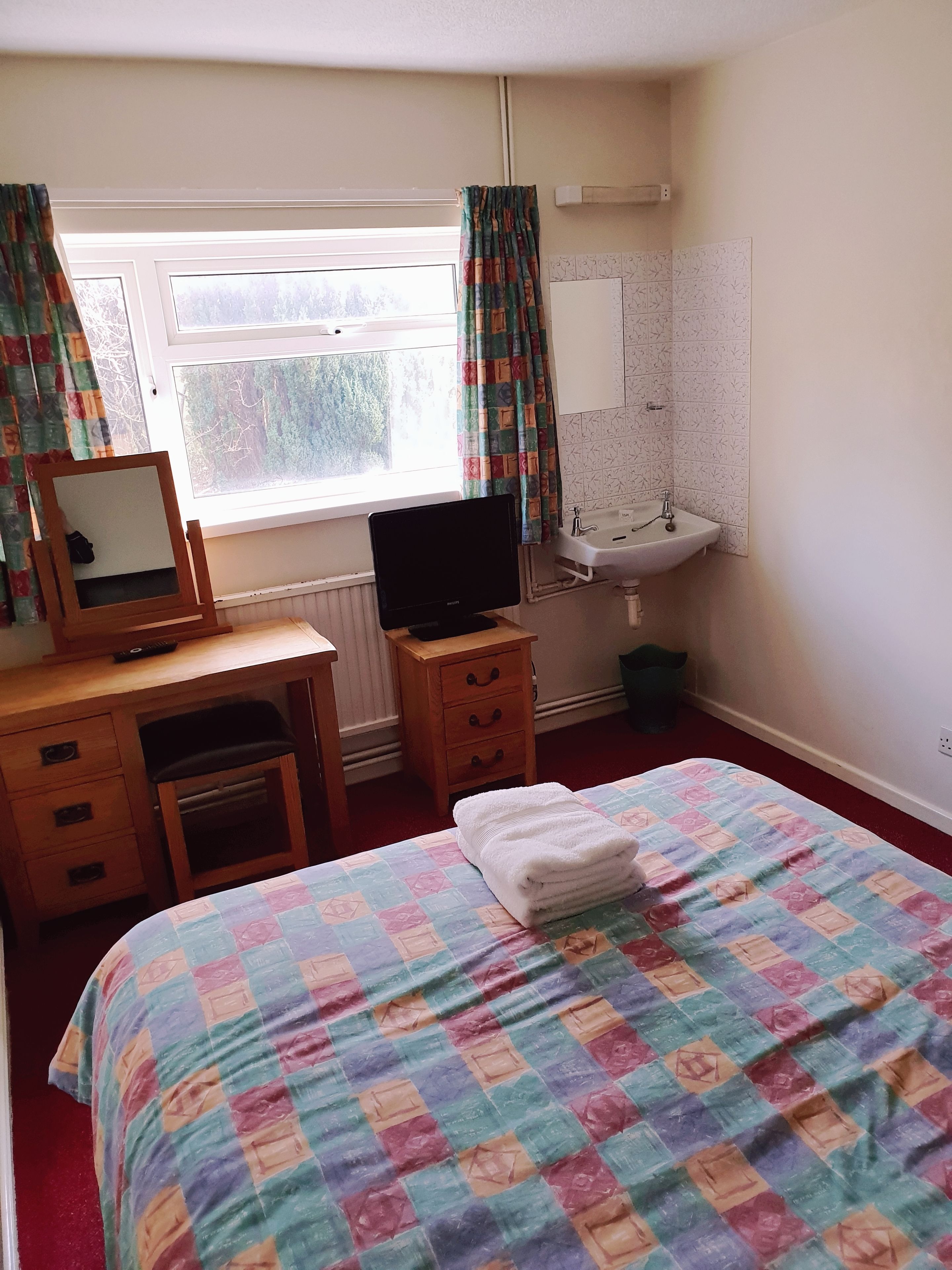 A bedroom with a bed, a desk with a computer, a window with curtains, a sink, and a mirror.