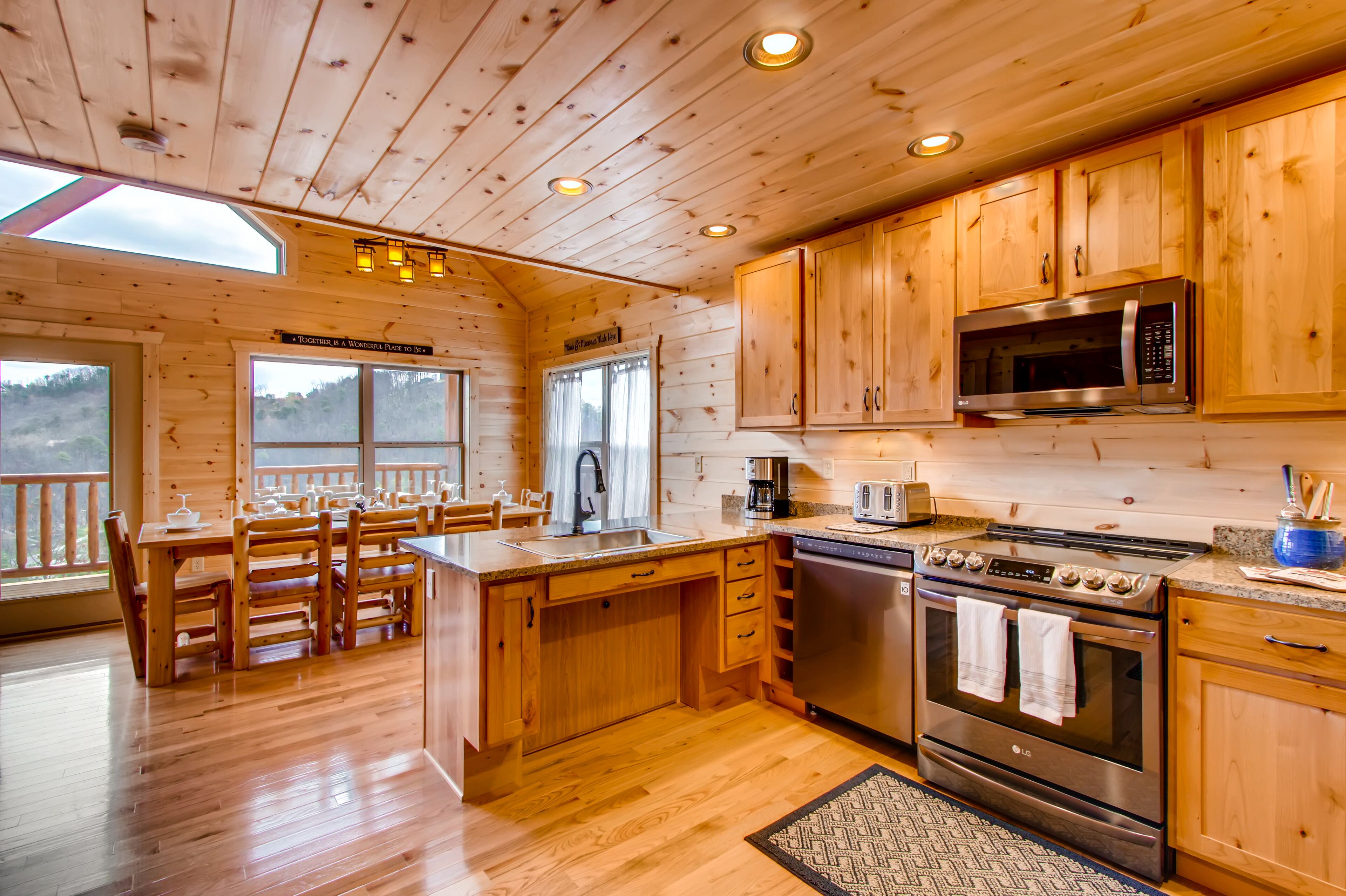 A wooden cabin kitchen with a dining area, wooden cabinets, stainless steel appliances, and a large window.