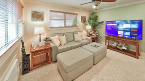 Ein Wohnzimmer mit einer Couch, einem Hocker, Holztischen, einem Fernseher und einem Deckenventilator.