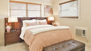Ein Schlafzimmer mit einem Bett, zwei Nachttischen, einem Schreibtischchen, einem Deckenventilator und einem Fenster mit Jalousien.