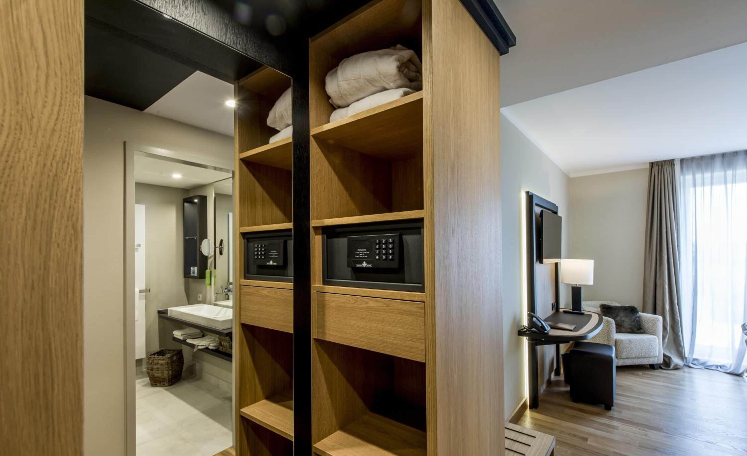 A modern hotel room with a wooden wardrobe, a vanity with a mirror, a small table with a lamp, and a sofa.