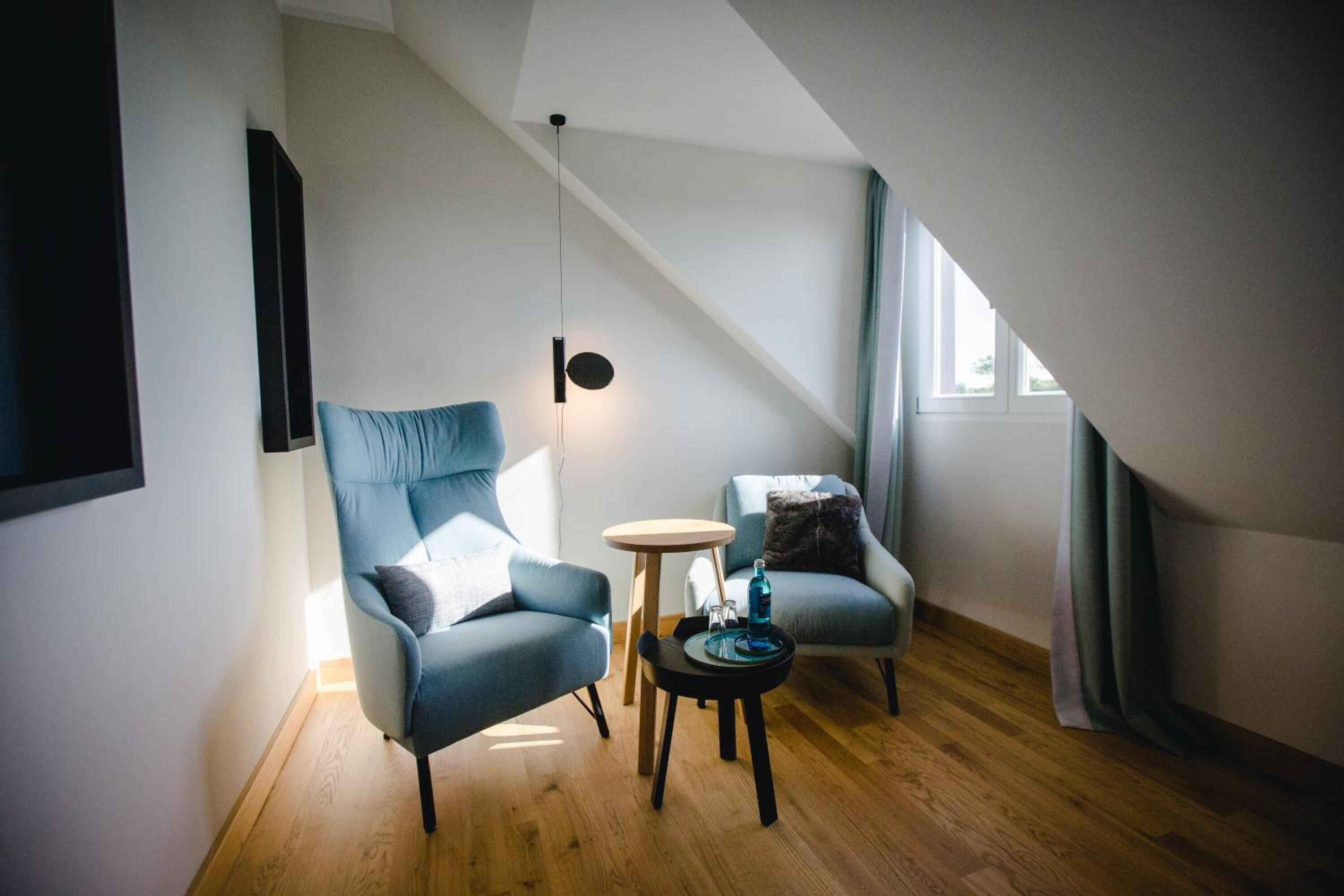 Een moderne loft slaapkamer met een schuine plafond, twee zithoeken met blauwe stoelen, een ronde houten tafel en een wandlamp.