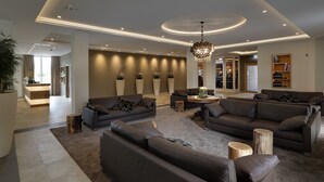 A modern living room with sofas, a coffee table, and a chandelier. There are large windows and a reception area in the background. - Hofhotel Grothues-Potthoff (Senden)