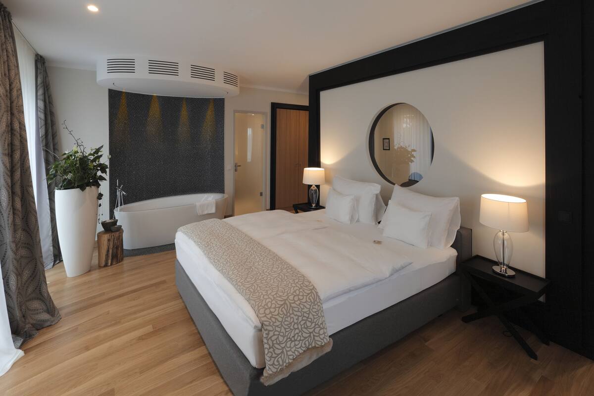 A modern hotel room with a large bed, a sitting area with a bathtub, and a large mirror on the wall.