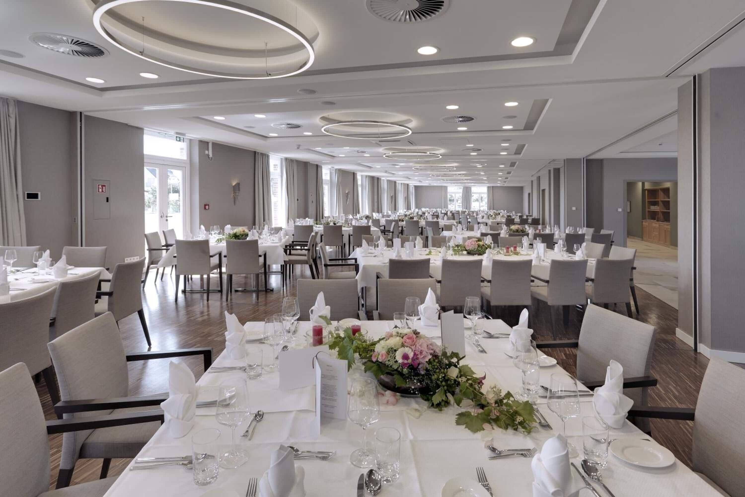 Un amplio salón de banquetes con mesas redondas dispuestas para el servicio de comidas, con manteles blancos y centros de mesa florales.